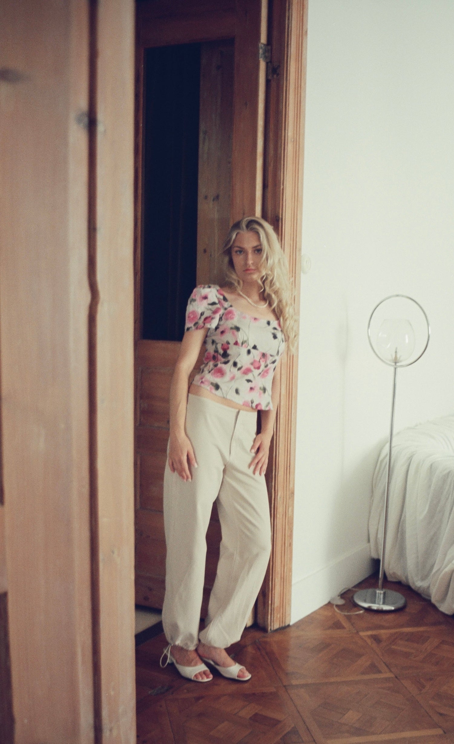 A woman stands in a doorway wearing a short-sleeve floral print top and cream-colored trousers, posing in a room with a wooden floor and a white bed in the background.